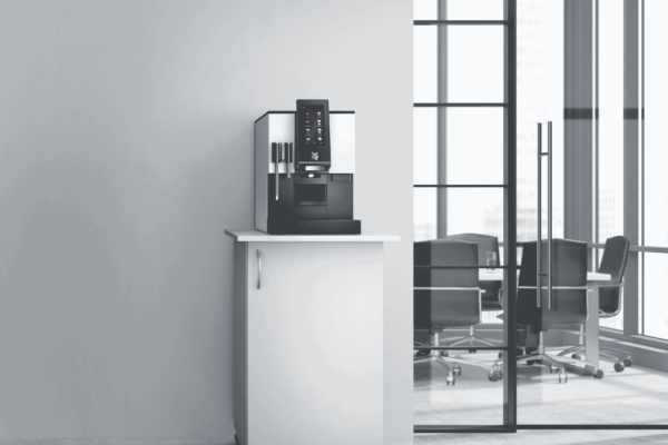 Nowoczesny ekspres do kawy WMF 1100 S stoi na białej szafce na kółkach, ustawionej przy ścianie w biurowym wnętrzu. W tle widoczne są przeszklone drzwi i sala konferencyjna z czarnymi krzesłami i dużymi oknami wpuszczającymi naturalne światło.