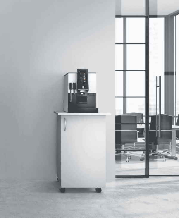 Nowoczesny ekspres do kawy WMF 1100 S stoi na białej szafce na kółkach, ustawionej przy ścianie w biurowym wnętrzu. W tle widoczne są przeszklone drzwi i sala konferencyjna z czarnymi krzesłami i dużymi oknami wpuszczającymi naturalne światło.