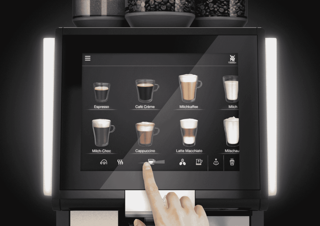 Zbliżenie na dotykowy wyświetlacz ekspresu do kawy WMF 1500 S+, na którym widoczne są różne opcje napojów, takie jak espresso, cappuccino, latte macchiato i inne. Palec osoby dotyka ekranu, wybierając rozmiar porcji kawy. Na górze urządzenia widoczne są pojemniki z ziarnami kawy. Całość utrzymana w nowoczesnym, eleganckim stylu z podświetleniem LED.